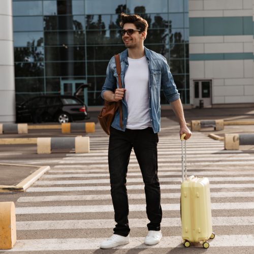 Brunette handsome man in white t-shirt and black pants walks on crosswalk. Cool guy in denim jacket and sunglasses holds brown backpack and yellow suitcase.