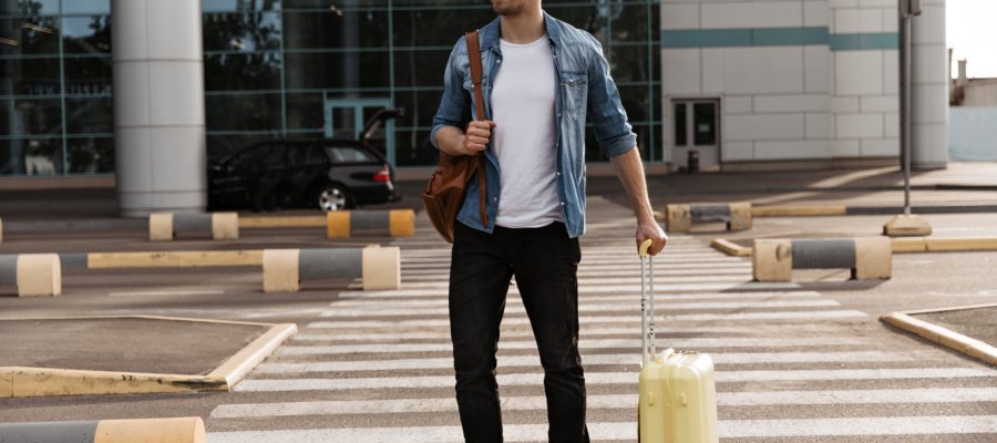 Brunette handsome man in white t-shirt and black pants walks on crosswalk. Cool guy in denim jacket and sunglasses holds brown backpack and yellow suitcase.